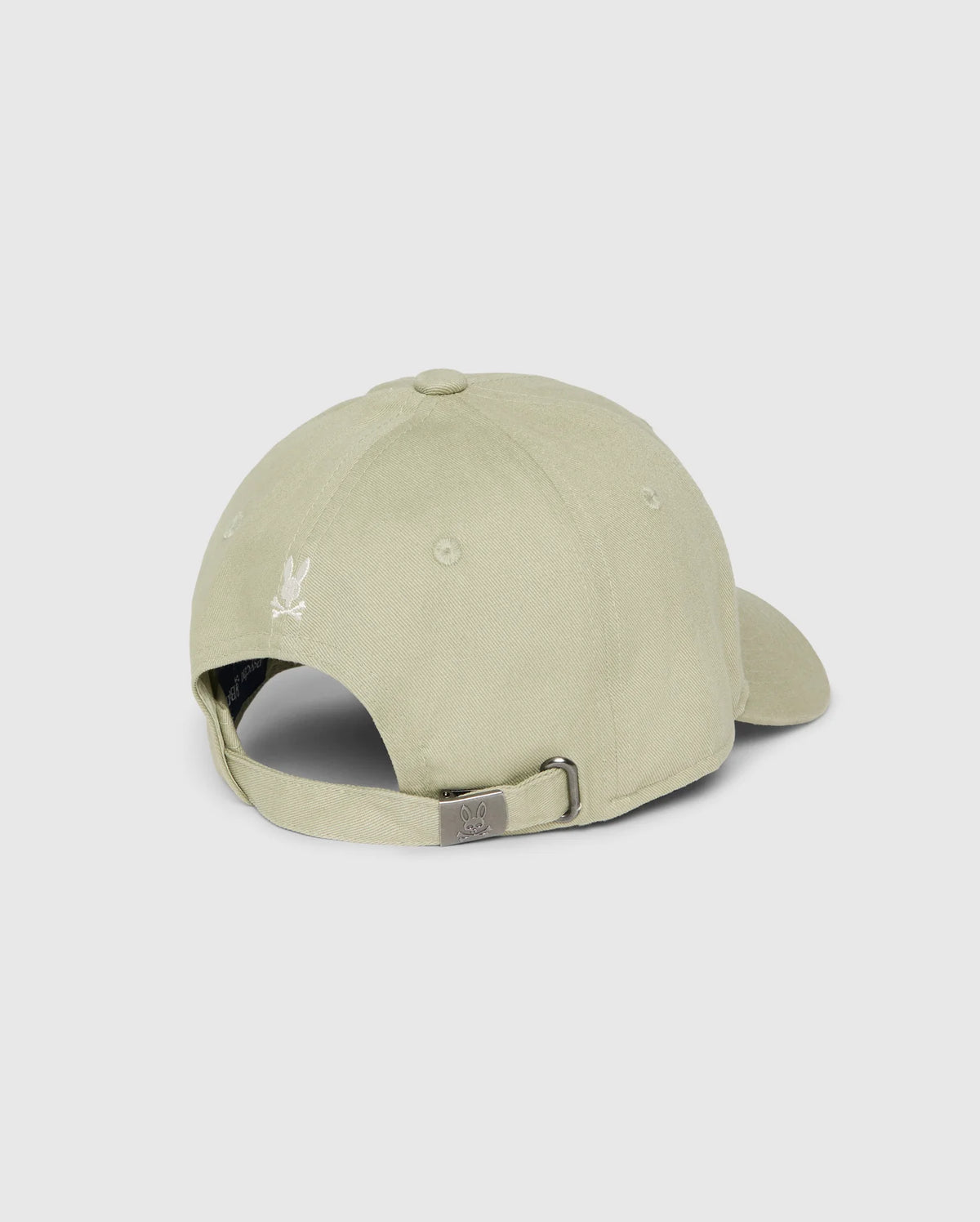 A light beige Psycho Bunny MENS KAYDEN BASEBALL CAP - B6A677C200 is shown from the back on a plain white background. The cap features an adjustable strap with a silver buckle. The back of the cap has ventilation eyelets and a small embroidered logo of a white dove near the top.