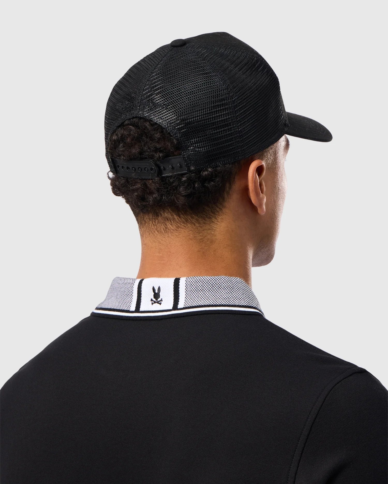 A man with short curly hair is seen from behind, wearing the MENS MALTON TRUCKER CAP - B6A797D200 by Psycho Bunny, a black trucker cap featuring mesh ventilation, paired with a 100% cotton polo shirt. The polo shirt has a white collar adorned with black and white stripes and features a small rabbit logo on the back. The background is plain light grey.
