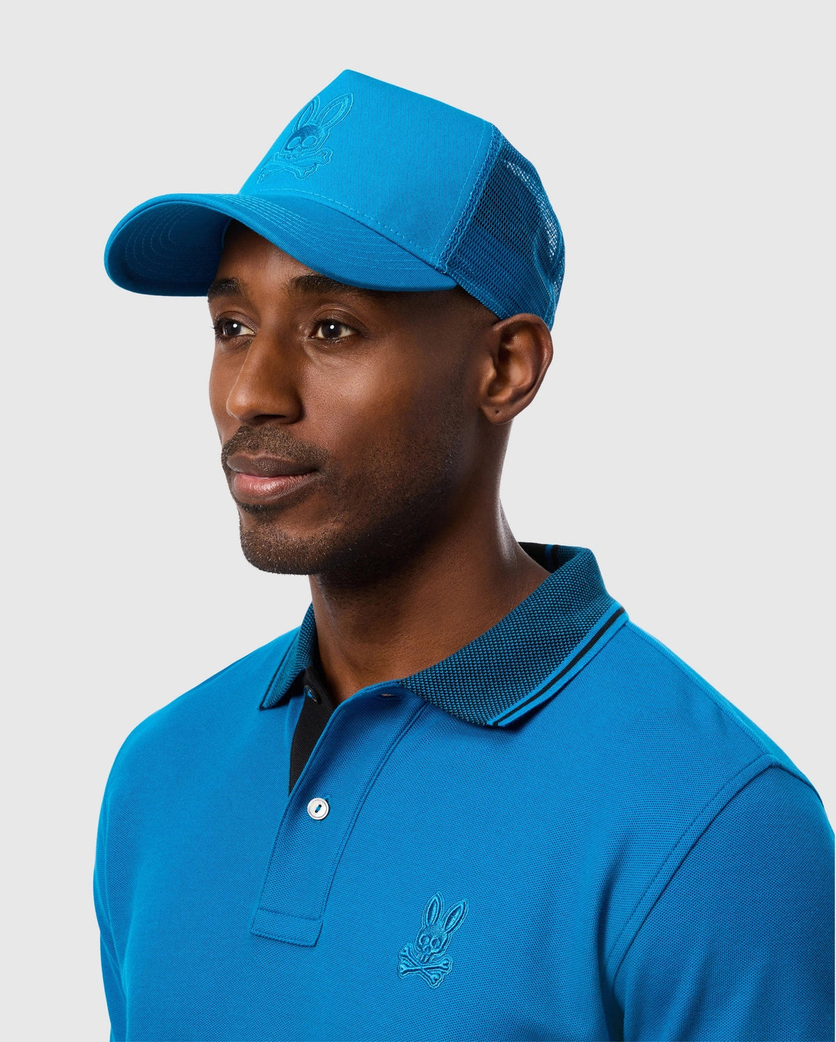 A man wearing the Psycho Bunny MENS MALTON TRUCKER CAP - B6A797D200, featuring mesh ventilation, along with a matching blue polo shirt that has an embroidered logo on the chest, looks to the right. The background is plain and light gray.