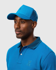 A man wearing the Psycho Bunny MENS MALTON TRUCKER CAP - B6A797D200, featuring mesh ventilation, along with a matching blue polo shirt that has an embroidered logo on the chest, looks to the right. The background is plain and light gray.