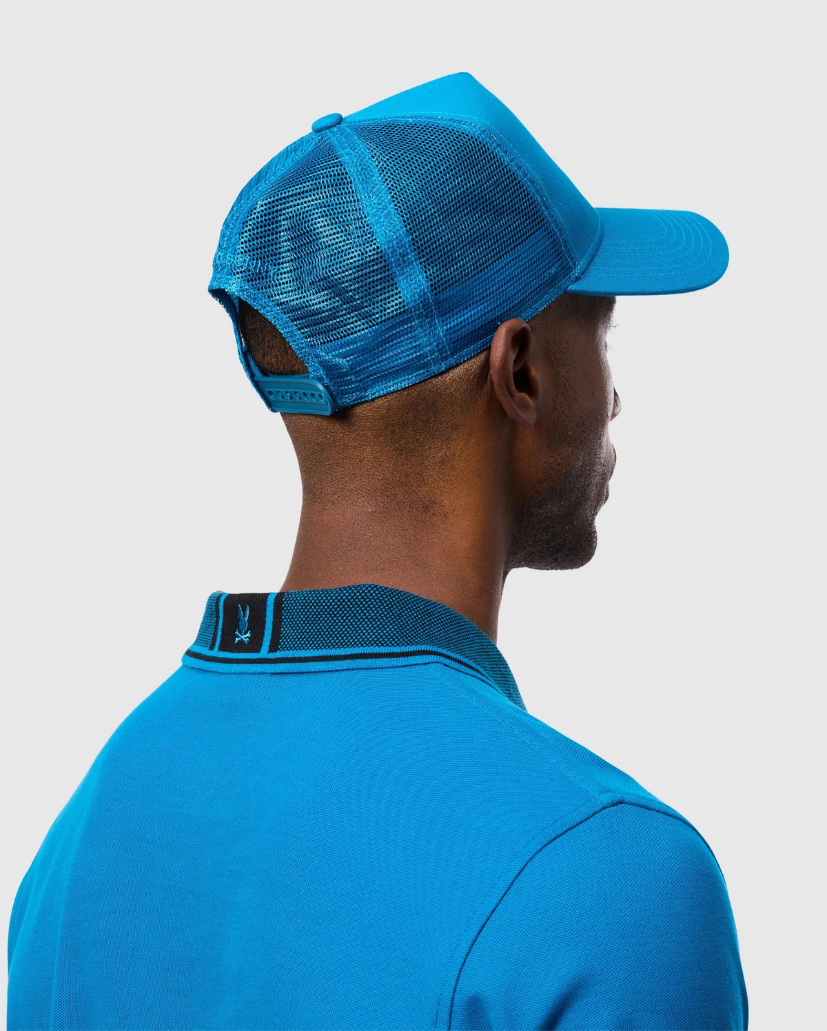 A person wearing a blue Psycho Bunny MENS MALTON TRUCKER CAP - B6A797D200 with excellent ventilation and a blue 100% cotton polo shirt is shown from the back. The cap features an adjustable strap, and the shirt has a logo on the back of the collar. The background is plain white.