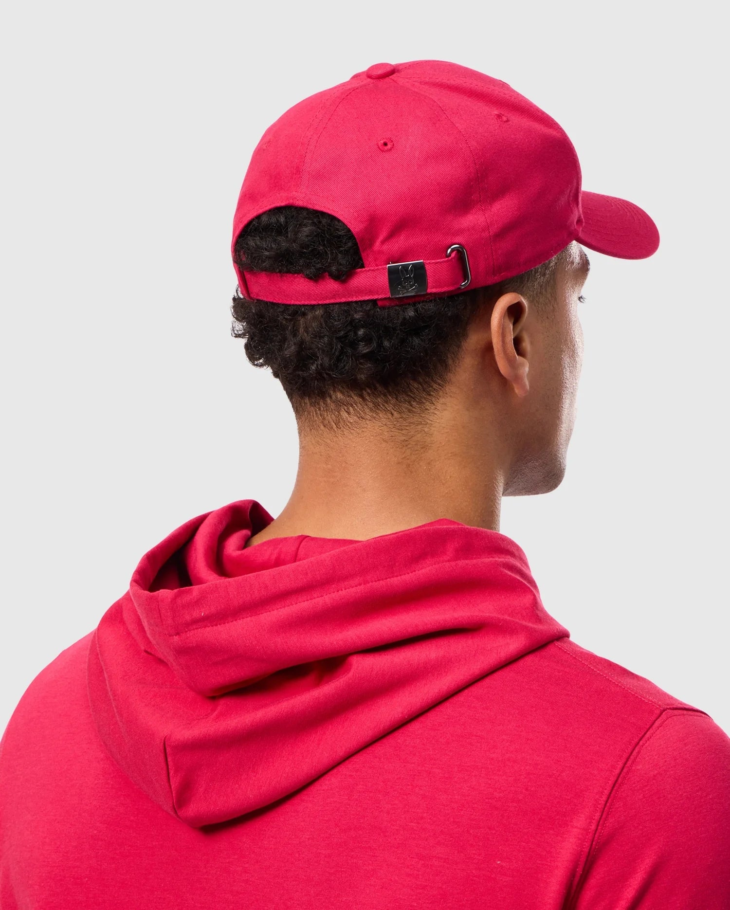 A person wearing a red hooded sweatshirt and a Psycho Bunny Men's Walter Baseball Cap (B6A800D200) is shown from behind. The hat, crafted from durable and comfortable 100% cotton, features an adjustable strap, set against a plain gray background.