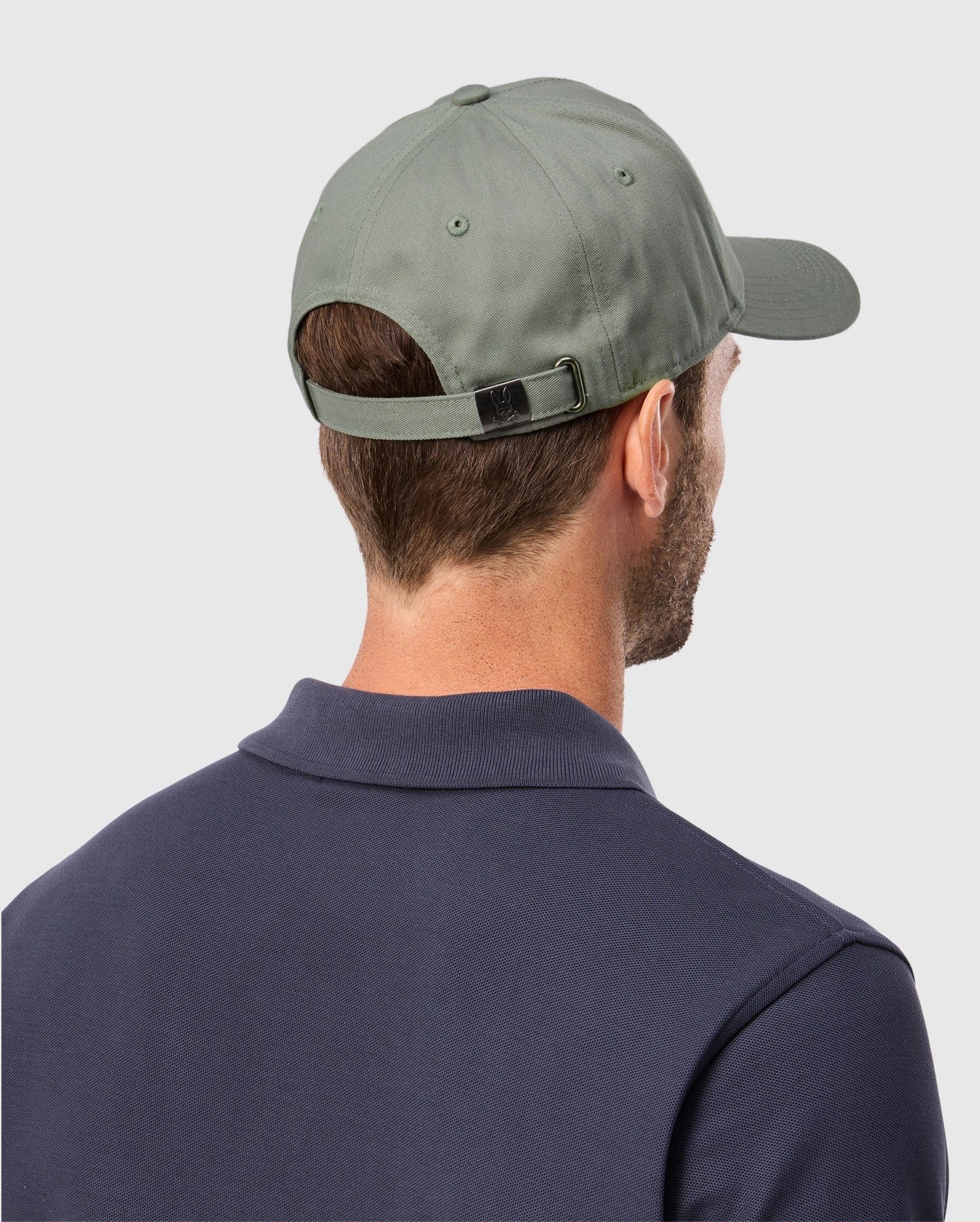 A person is shown from the back wearing a Psycho Bunny Men's Classic Baseball Cap (B6A816B200) in green and a dark polo shirt. The cap's strap highlights its solid pattern, set against a light gray background.