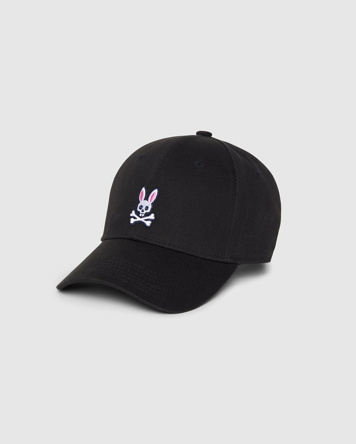 A Psycho Bunny MENS CLASSIC BASEBALL CAP - B6A816A1HT in black featuring a small embroidered pink and white bunny above crossed bones on the front. Made from durable cotton twill, it has a curved brim and six panels with eyelets for ventilation. The background is plain and light gray.