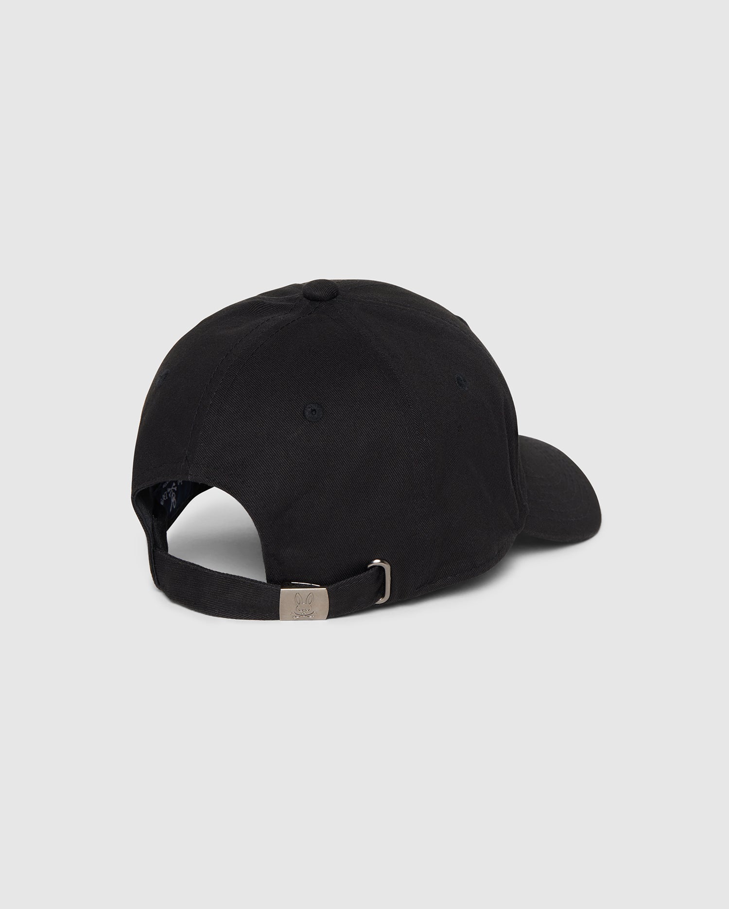 A Psycho Bunny MENS CLASSIC BASEBALL CAP - B6A816A1HT is shown from the back. The black cotton twill cap features an adjustable strap with a metal buckle and a small round vent hole on the side. The background is a plain grey.