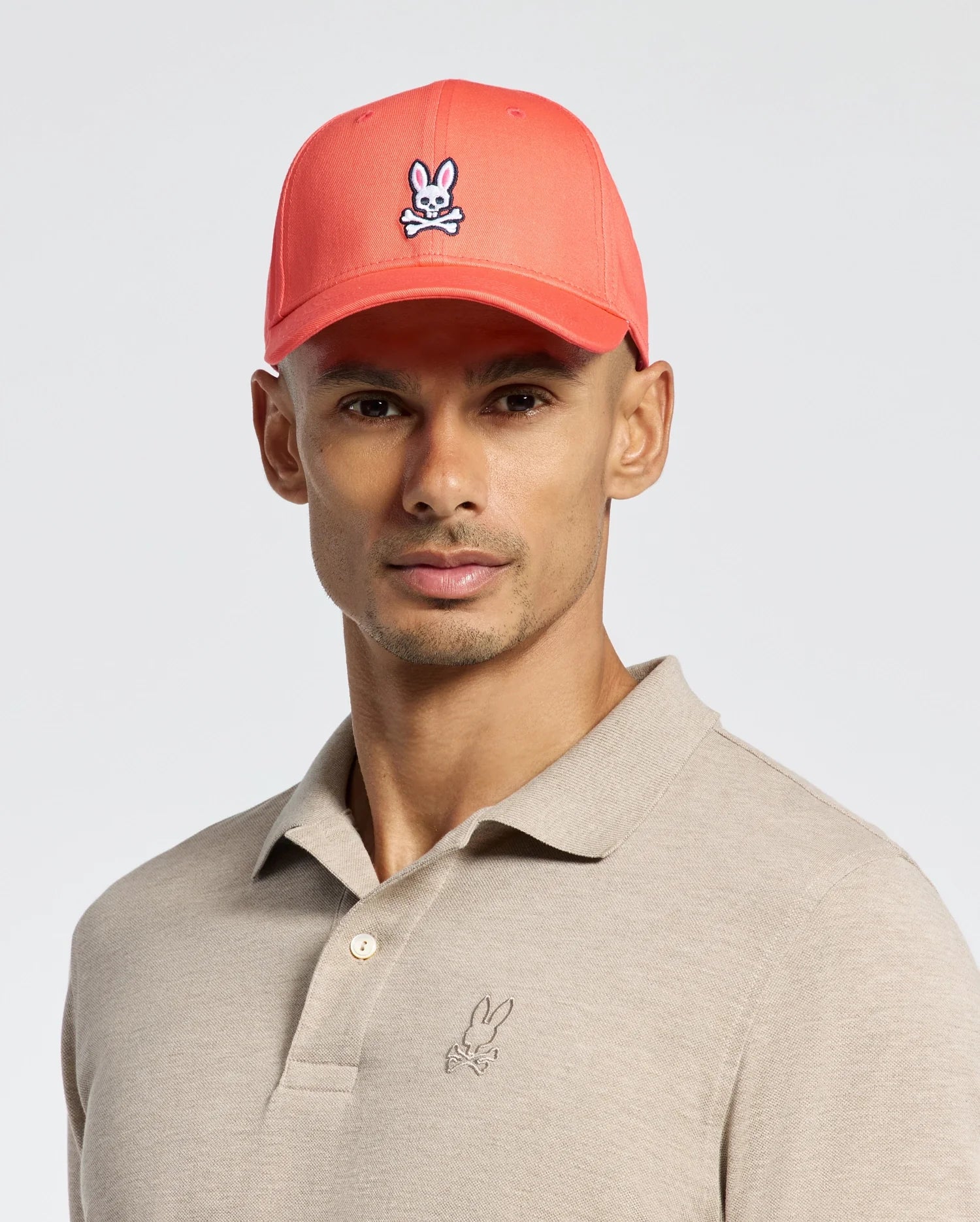 A man wears a Psycho Bunny MENS CLASSIC BASEBALL CAP - B6A816F200 with an embroidered rabbit logo in coral cotton twill. His beige polo also features this logo, and the adjustable cap fits perfectly against the plain light gray background.