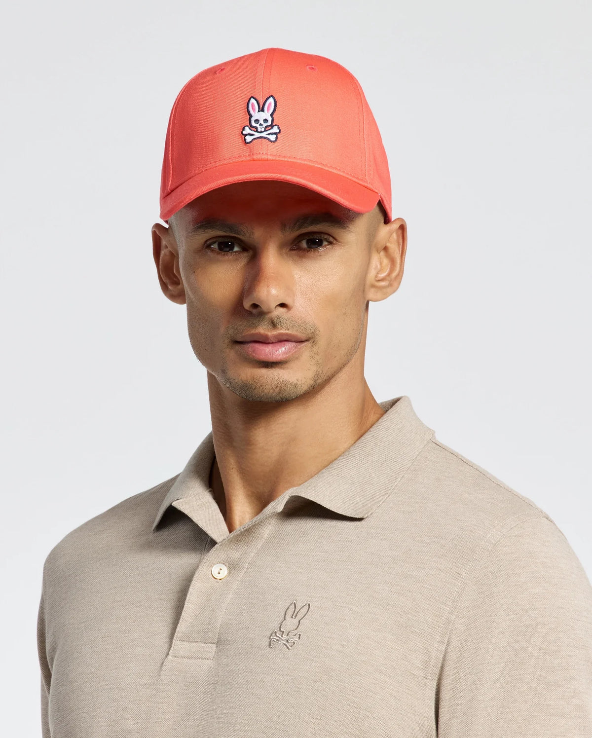 A man wears a Psycho Bunny MENS CLASSIC BASEBALL CAP - B6A816F200 with an embroidered rabbit logo in coral cotton twill. His beige polo also features this logo, and the adjustable cap fits perfectly against the plain light gray background.