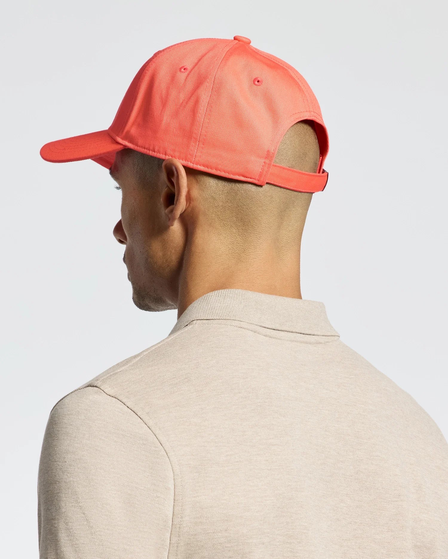A person is shown from the side and back wearing a beige shirt and Psycho Bunny's MENS CLASSIC BASEBALL CAP in bright coral (B6A816F200), featuring a visible adjustable fit strap. The background is plain and light-colored.