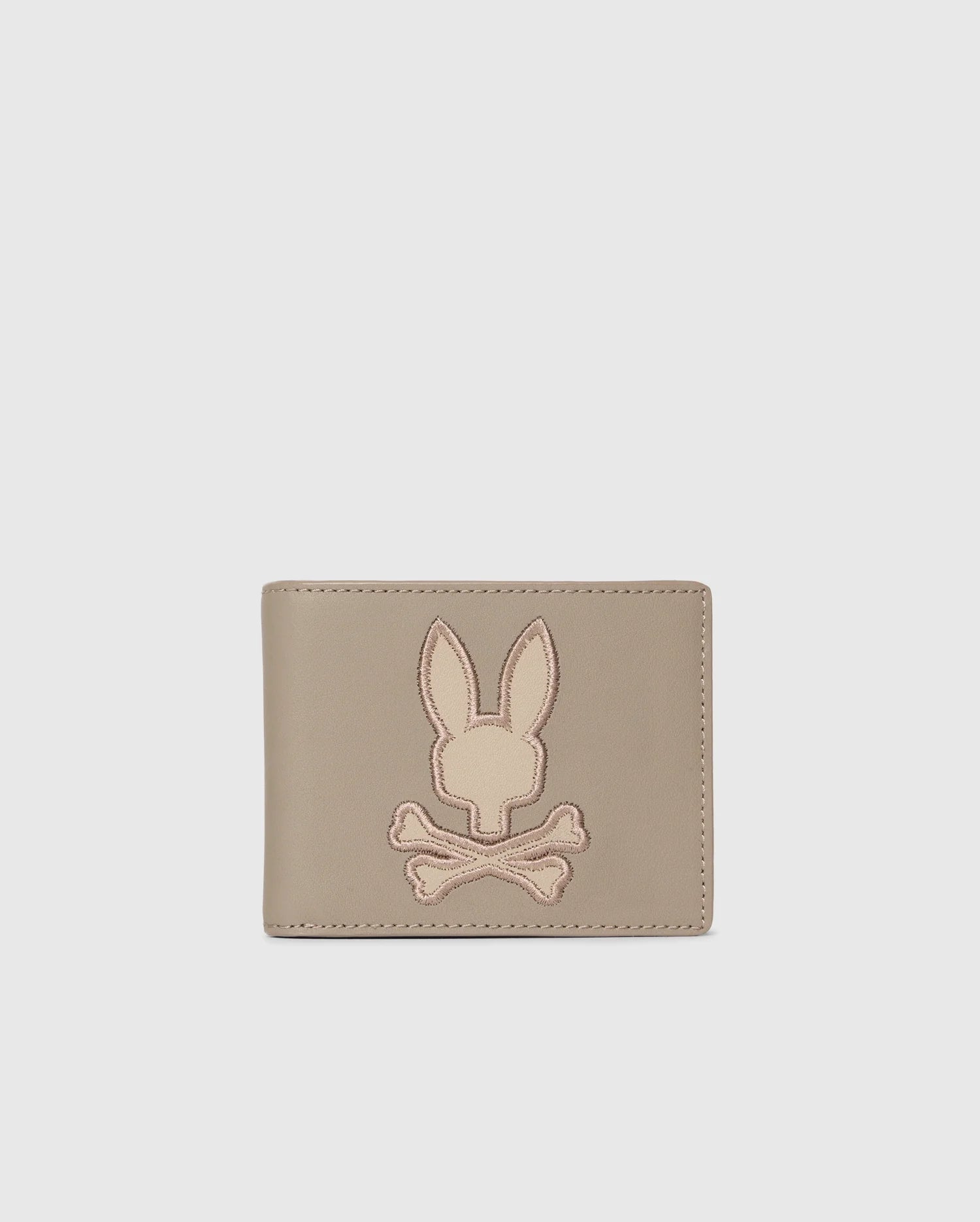 The Psycho Bunny MENS LEATHER WALLET - B6A868D200, featured on a plain background, displays an embossed rabbit's head with crossed bones. This beige wallet offers a minimalist and stylish design for those who value refined craftsmanship.