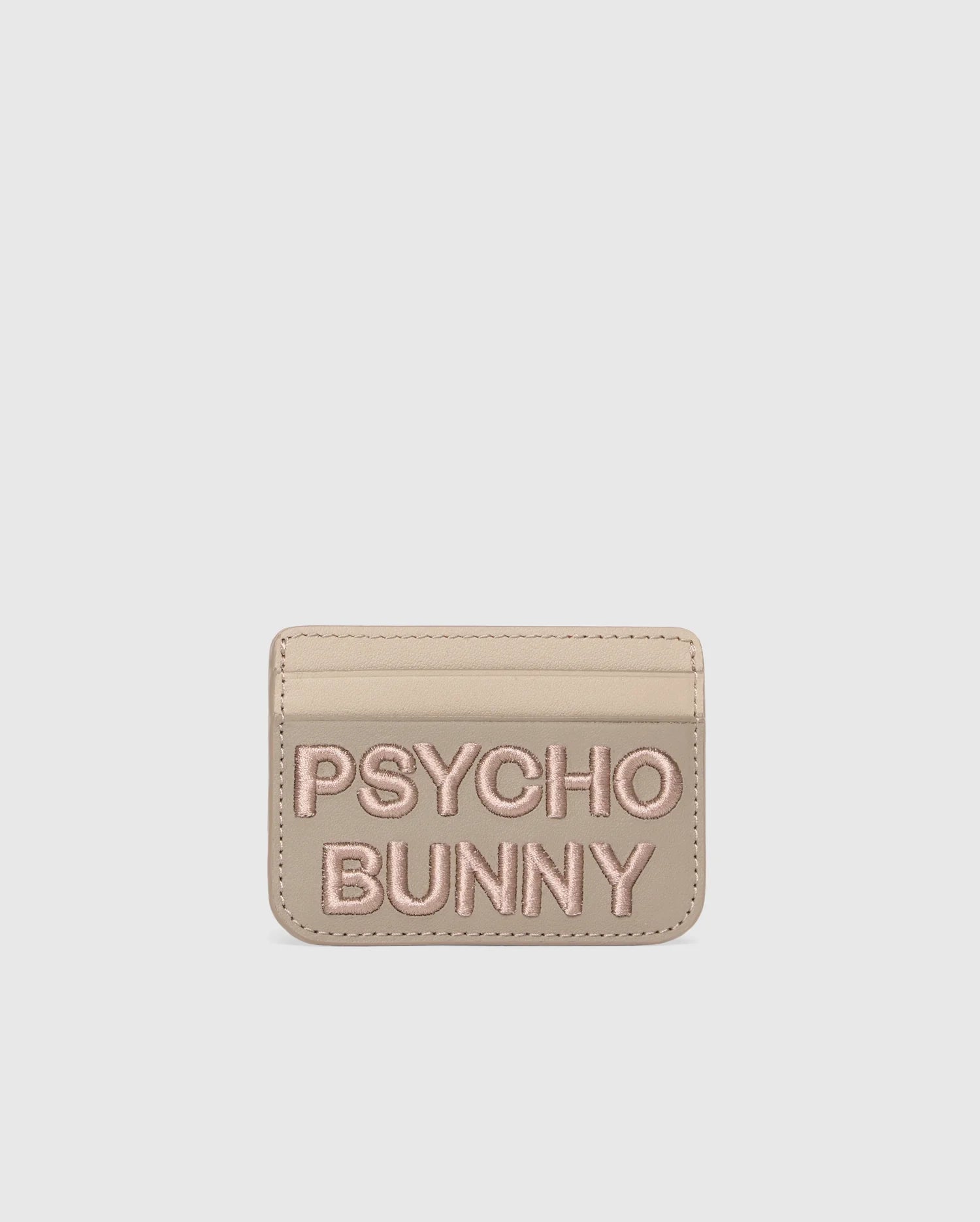 A premium beige leather cardholder features "PSYCHO BUNNY" embroidered in pink on the front. The minimalist MENS LEATHER CARD CASE - B6A869D200 by Psycho Bunny offers style and simplicity, set against a plain white background.