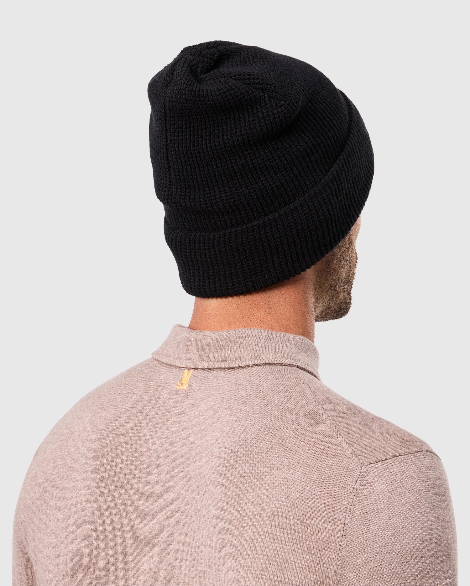 Rear view of a person wearing a cozy beige sweater and the Psycho Bunny MENS WAFFLE BEANIE - B6A873D200 in black, standing against a plain light gray background.