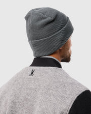 A person is depicted from behind wearing a Psycho Bunny MENS WAFFLE BEANIE - B6A873D200 in gray cotton, paired with a gray jacket that has a black collar and showcases a subtle black rabbit logo on the back. The setting is against a plain light gray background.