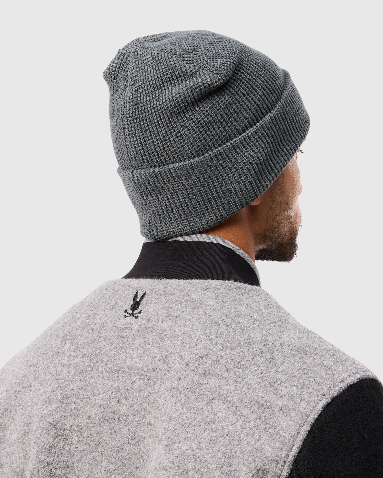 A person is depicted from behind wearing a Psycho Bunny MENS WAFFLE BEANIE - B6A873D200 in gray cotton, paired with a gray jacket that has a black collar and showcases a subtle black rabbit logo on the back. The setting is against a plain light gray background.
