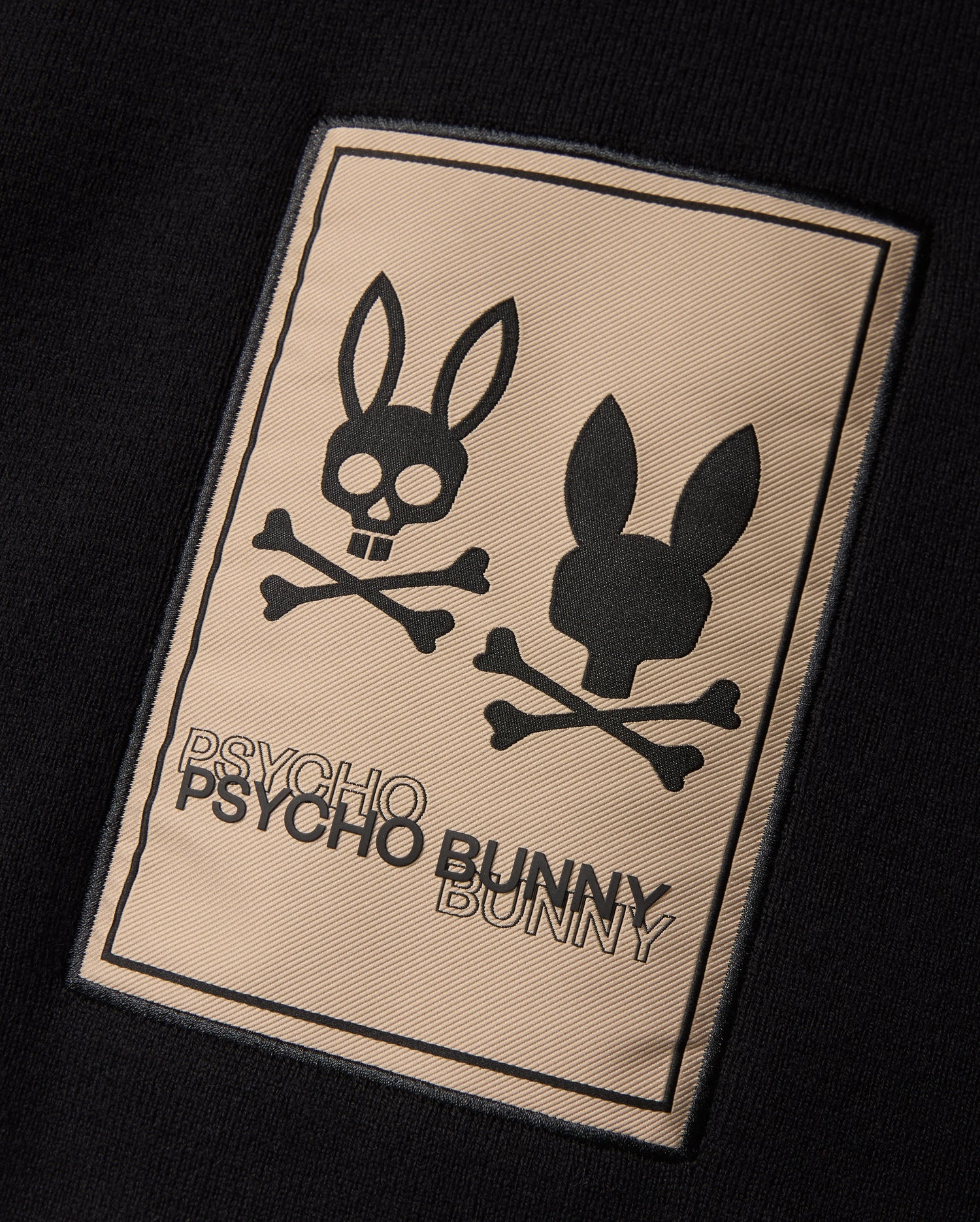 The MENS MONROE TWILL PATCH SCARF by Psycho Bunny features a luxurious black cotton wool blend. It boasts a rectangular patch embellished with two bunny head skull and crossbones designs. The brand name "Psycho Bunny" is elegantly emblazoned in bold lettering below, enhancing its edgy sophistication.