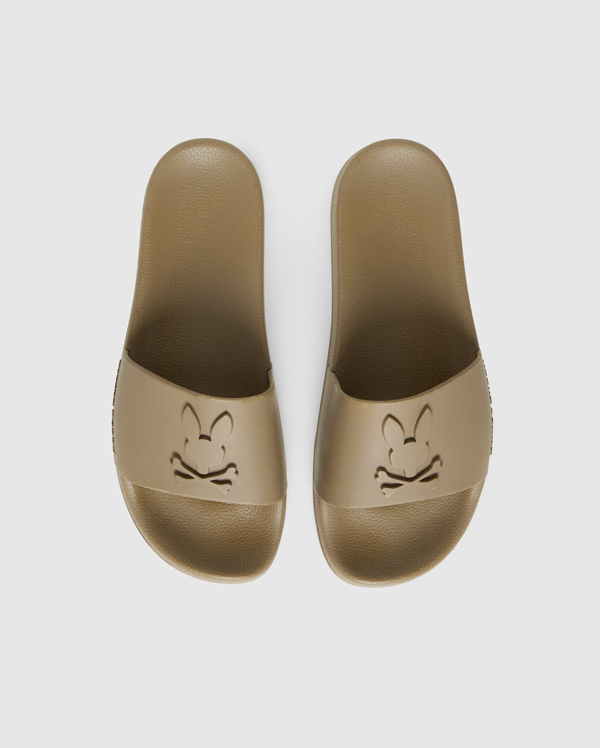 A pair of brown pool slides with an embossed cartoon rabbit face design on each top strap, viewed from above on a white background are the MENS LOGO SLIDES - B6A910U1FT by Psycho Bunny.