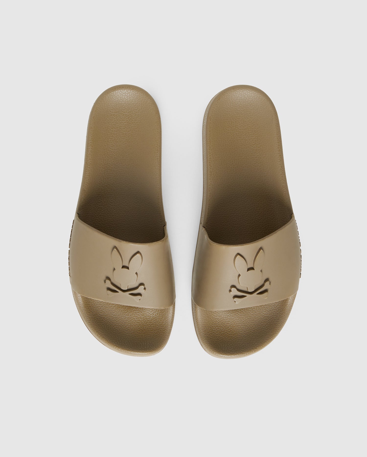 A pair of brown pool slides with an embossed cartoon rabbit face design on each top strap, viewed from above on a white background are the MENS LOGO SLIDES - B6A910U1FT by Psycho Bunny.