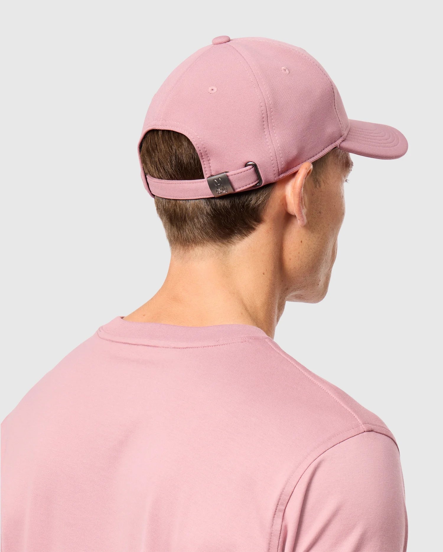 A person is facing away from the camera, wearing a pink MENS JAMES PREMIUM BASEBALL CAP by Psycho Bunny and a matching pink shirt. The cap, crafted from polyester spandex, features an adjustable strap with a metal buckle and showcases an embroidered Bunny design on the front. The background is plain gray.