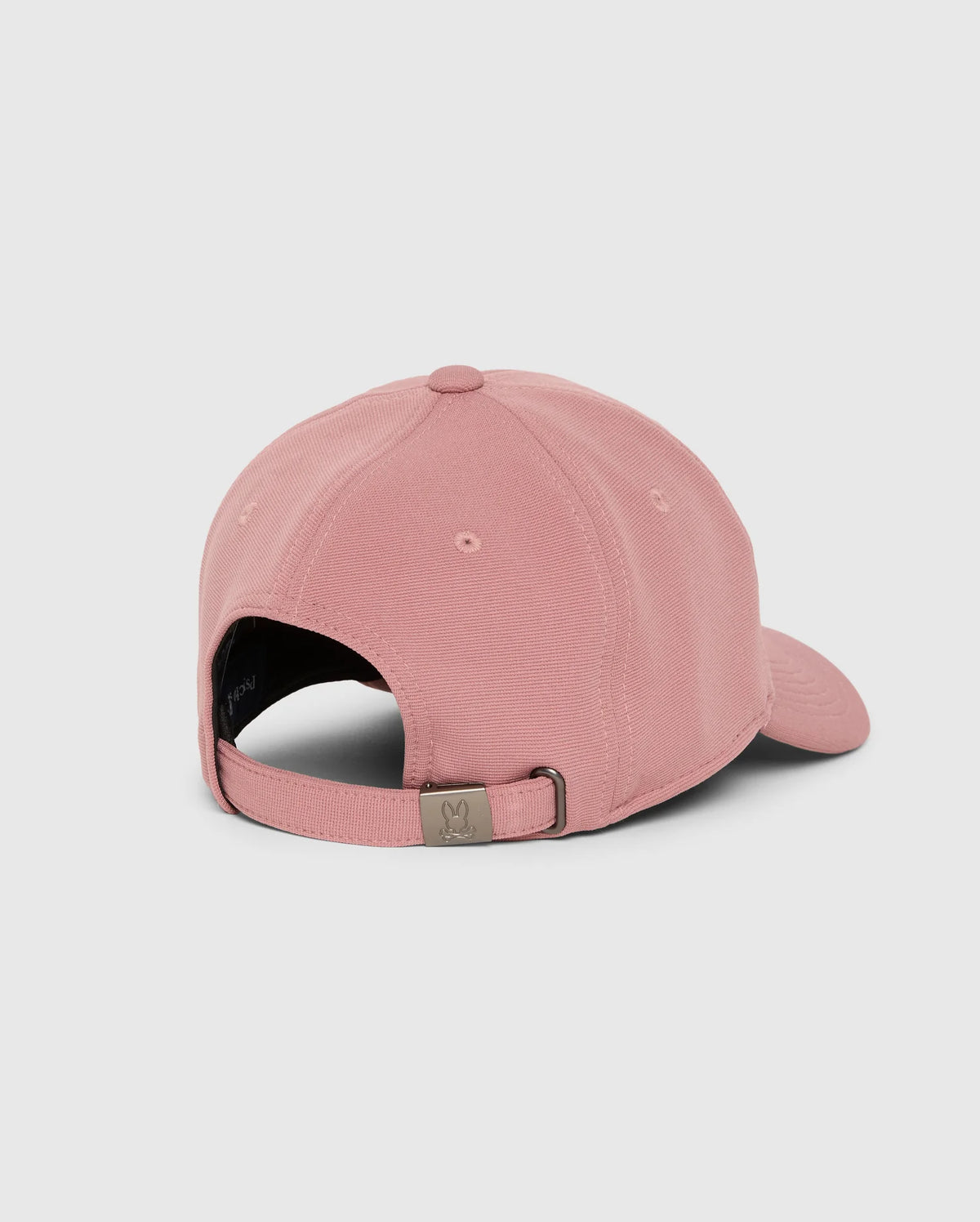 The MENS JAMES PREMIUM BASEBALL CAP - B6A953D200 by Psycho Bunny in pink is displayed from the back, highlighting its adjustable strap with a metal clasp adorned with a small symbol. Crafted from polyester spandex, this cap also features an embroidered Bunny detail. It is presented against a plain, light gray background.