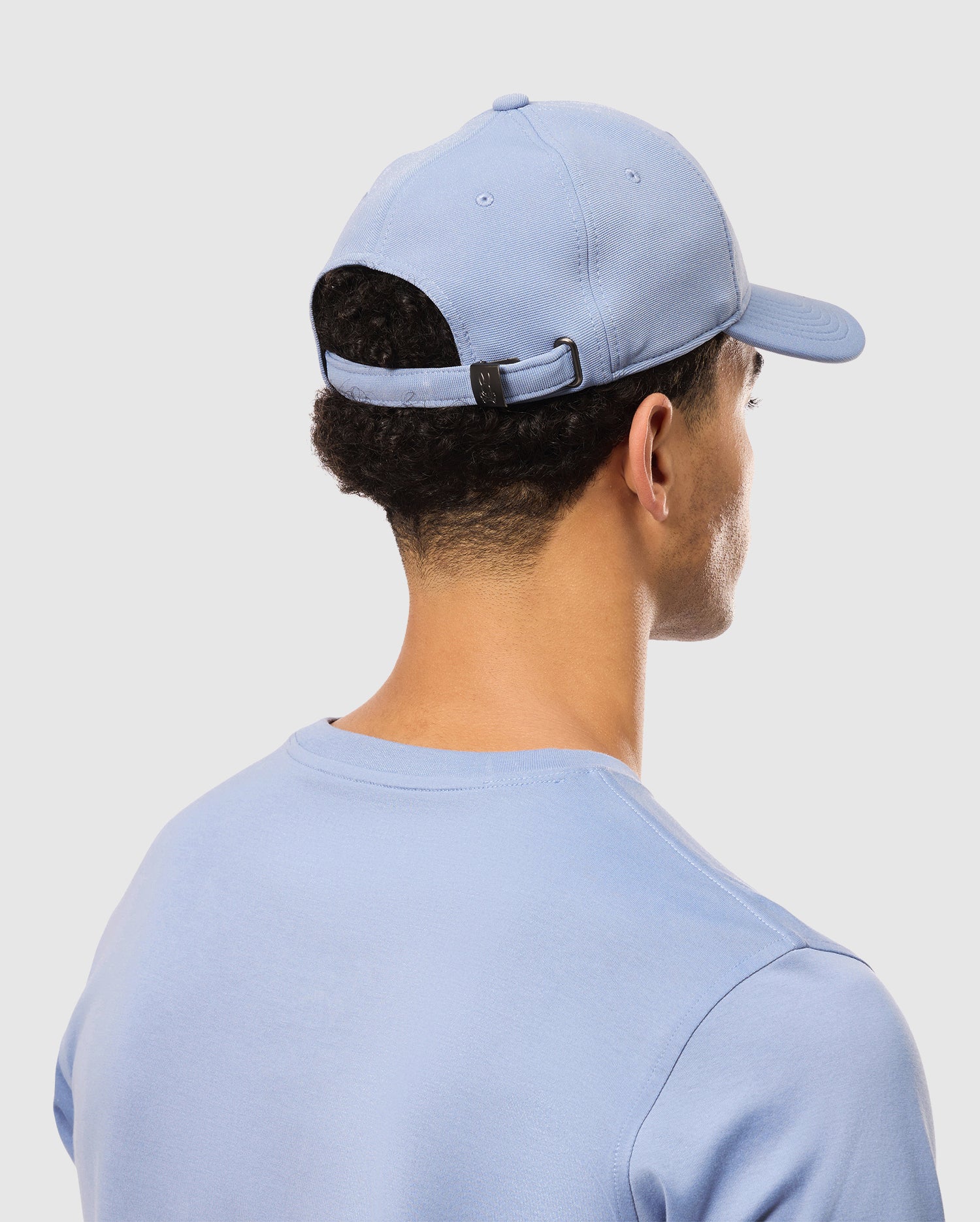 A person wears a Psycho Bunny MENS JAMES PREMIUM BASEBALL CAP (B6A953D200) in light blue with a matching shirt, viewed from behind. The knit twill cap includes an adjustable strap with a metal buckle, set against a plain light background.