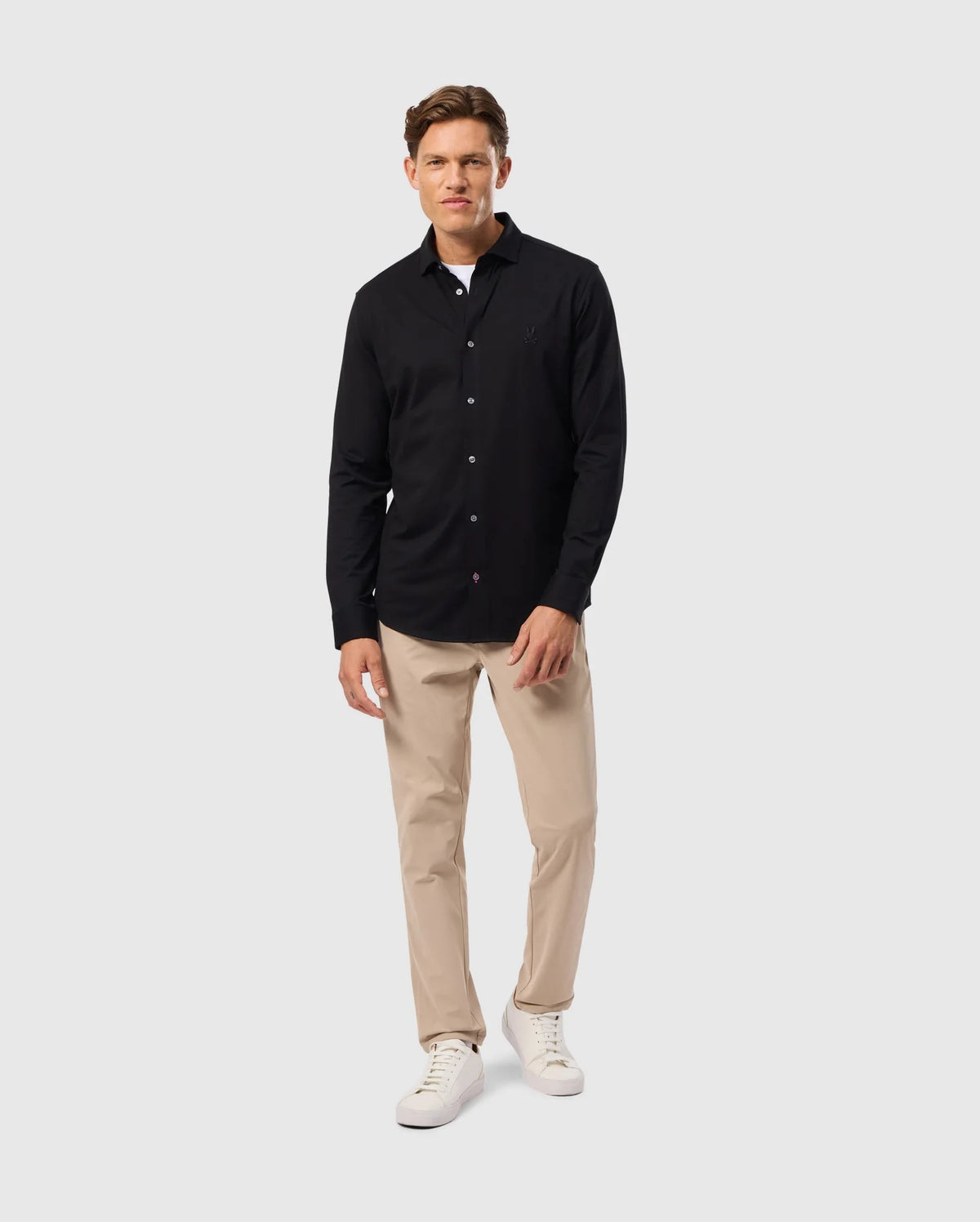 A man in a Psycho Bunny MENS ARTHUR JERSEY SHIRT stands against a light gray background. Wearing beige pants and white sneakers, he exudes a relaxed vibe with one hand in his pocket.