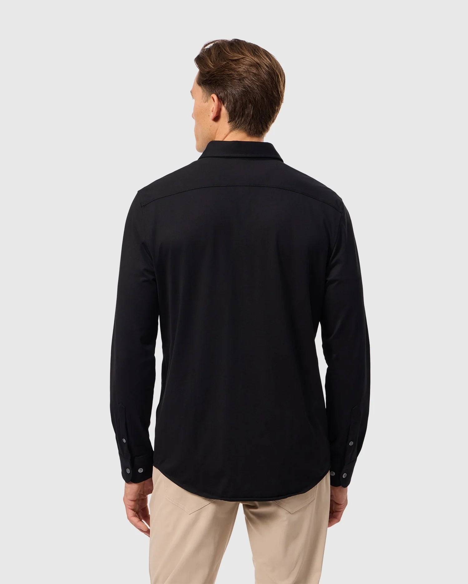 A man with short brown hair is turned away from the camera, highlighting a relaxed style in the Psycho Bunny MENS ARTHUR JERSEY SHIRT - B6C748D200, a long-sleeved black button-up crafted from Pima cotton, paired with beige pants, all set against a plain light gray backdrop.