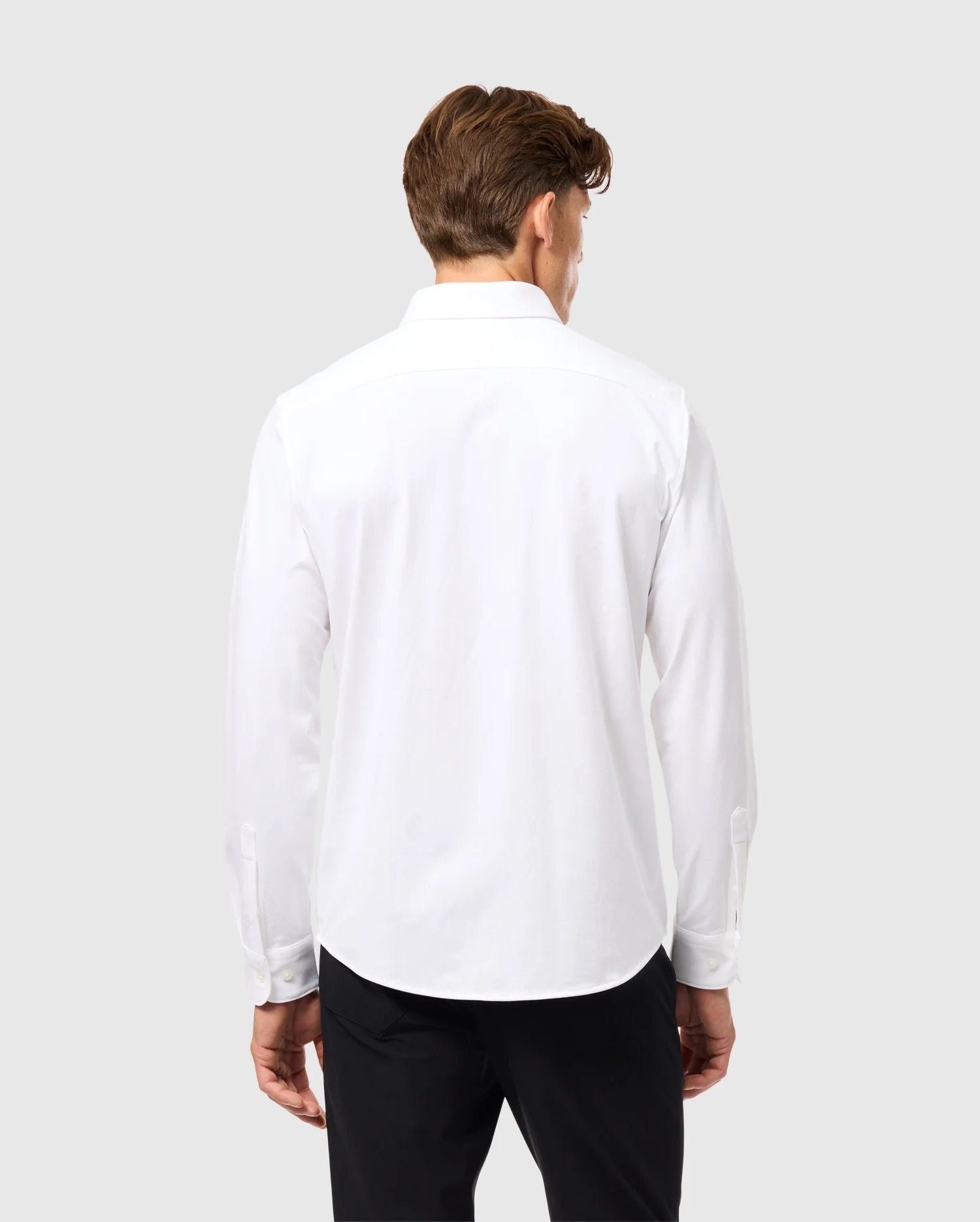A man with brown hair is facing away, wearing a Psycho Bunny MENS ARTHUR JERSEY SHIRT - B6C748D200 crafted from stretch Pima cotton, paired with black pants against a light gray background.