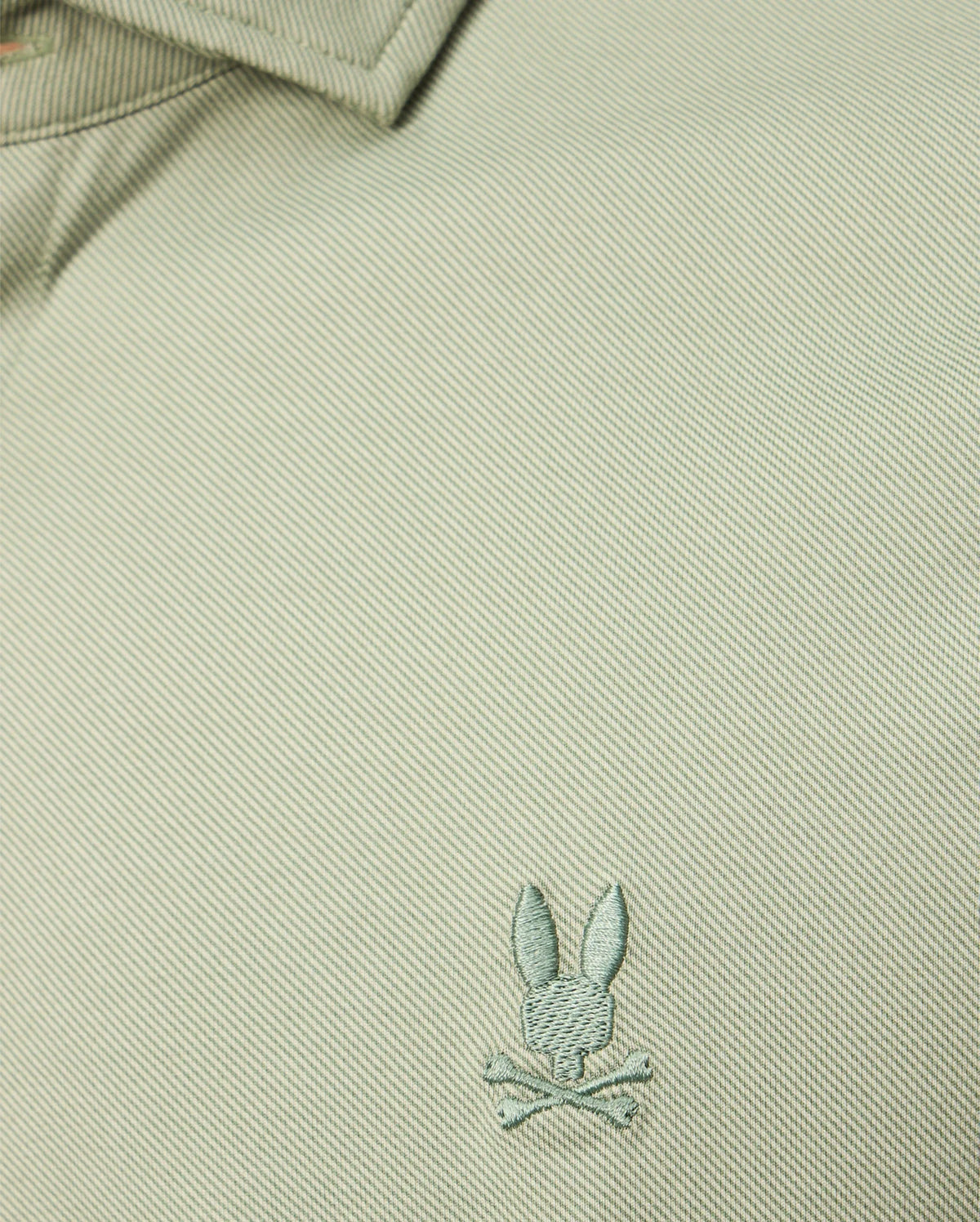 A close-up of the textured Pima cotton jersey from the Psycho Bunny MENS BRENDAN JERSEY SHIRT - B6C749D200 in light green highlights the embroidered chest Bunny logo, featuring a rabbit head with crossed bones beneath it. The design is elevated by subtle contrast piping that adds an extra touch of style.