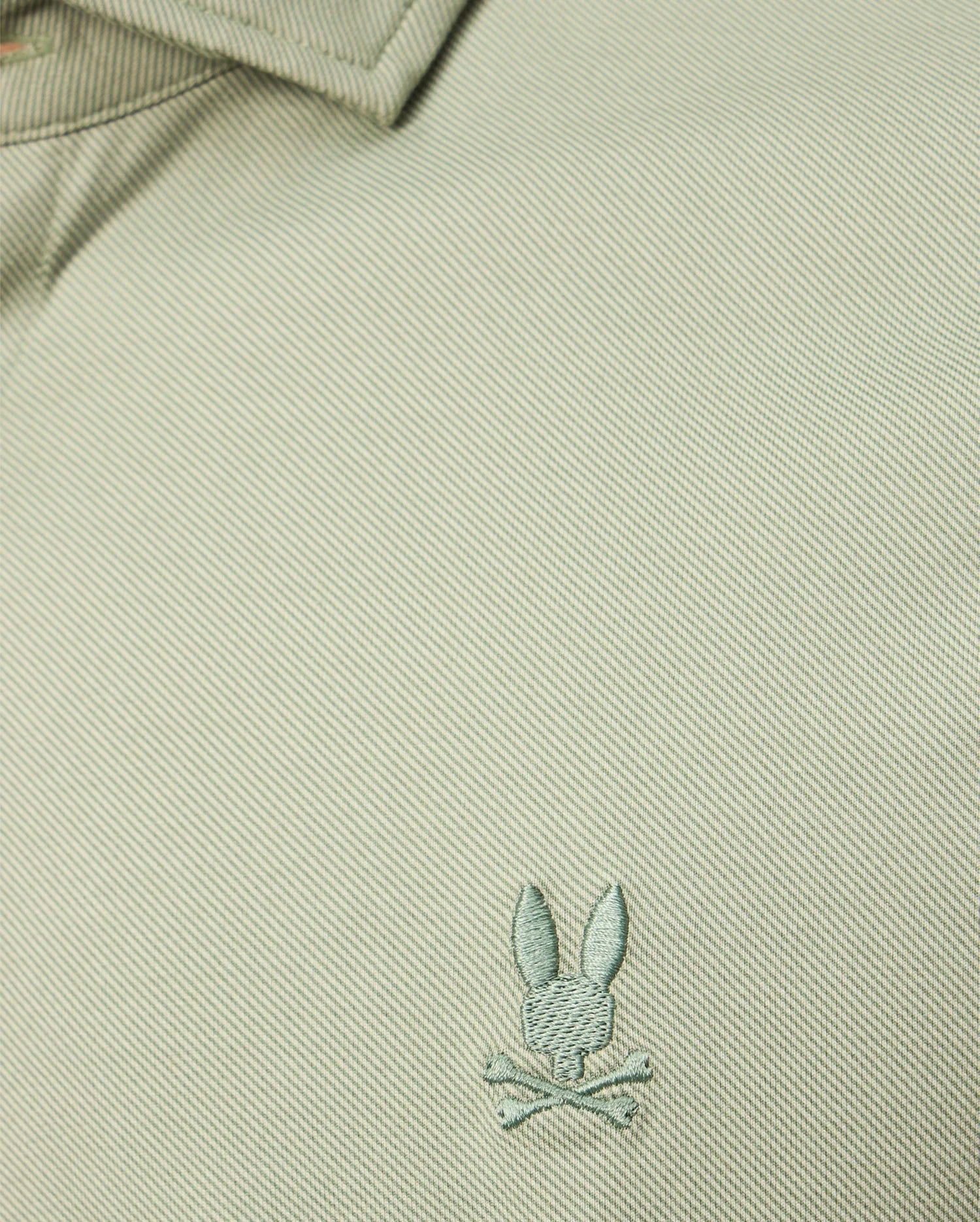 A close-up of the textured Pima cotton jersey from the Psycho Bunny MENS BRENDAN JERSEY SHIRT - B6C749D200 in light green highlights the embroidered chest Bunny logo, featuring a rabbit head with crossed bones beneath it. The design is elevated by subtle contrast piping that adds an extra touch of style.