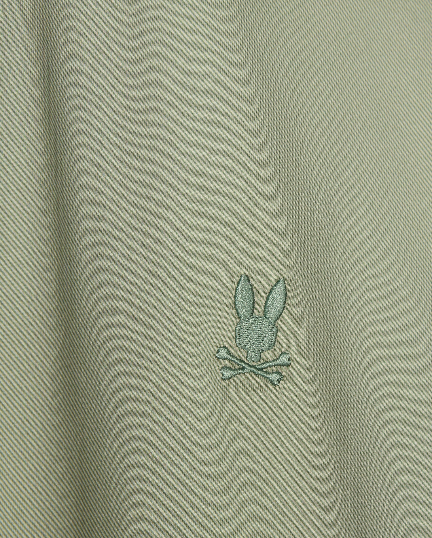 Close-up of the Psycho Bunny's Men's Brendan Jersey Shirt in light green Pima cotton, showcasing a subtle diagonal texture. The shirt features a small embroidered chest Bunny logo, depicting a rabbit head above two crossed bones, centered on the fabric.