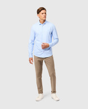 A person wearing the Psycho Bunny MENS BRENDAN JERSEY SHIRT in light blue Pima cotton and beige pants stands against a grey background. They are also sporting white sneakers and gazing slightly to the left.
