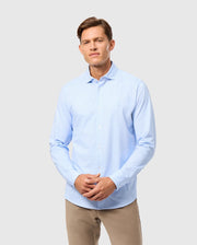 A person wearing a light blue MENS BRENDAN JERSEY SHIRT from Psycho Bunny, featuring an embroidered chest Bunny logo, and paired with beige pants stands against a plain background. The shirt is buttoned up with long sleeves. The individual has short hair and faces forward with a neutral expression.