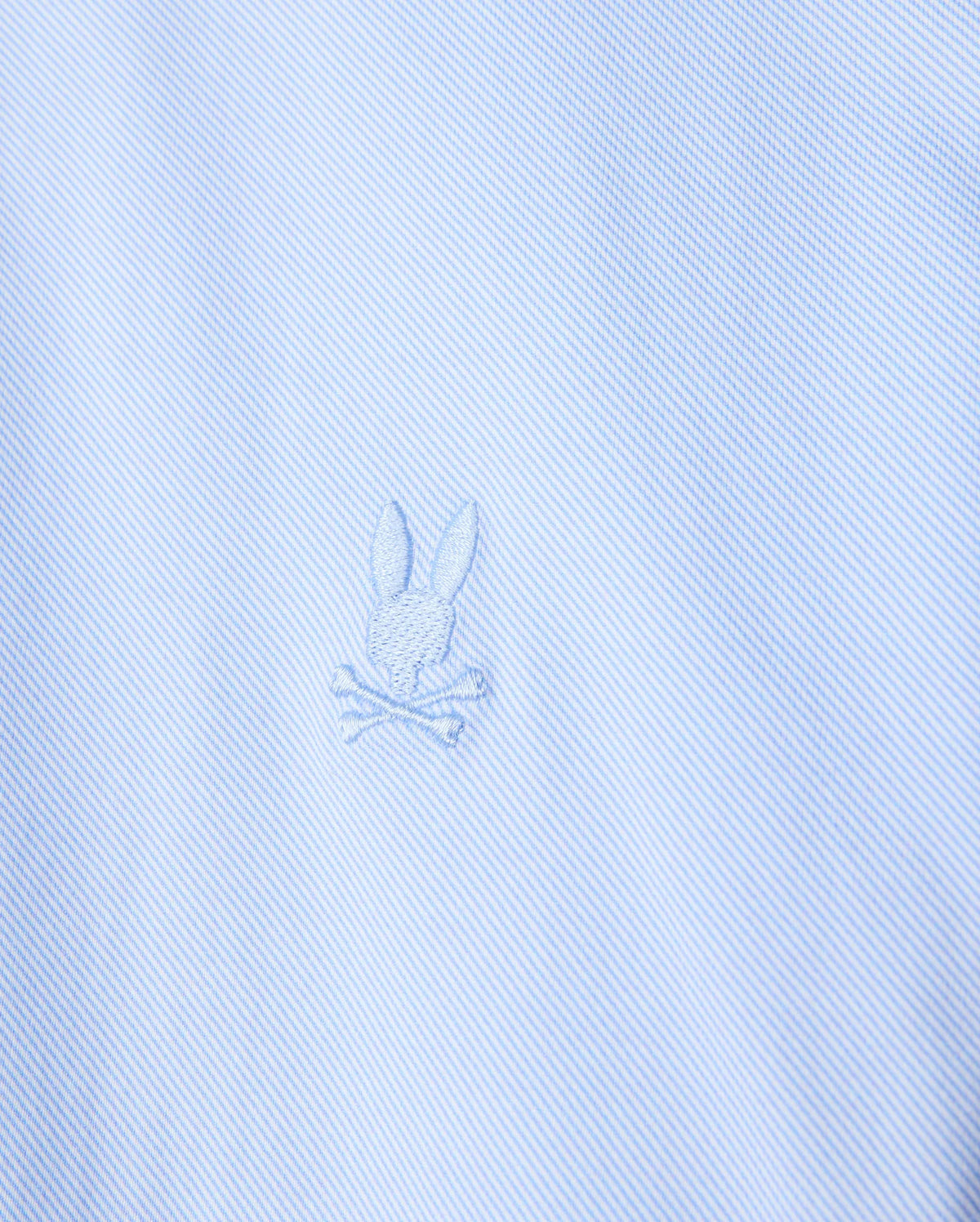 Close-up of the light blue stretch jersey fabric of the Psycho Bunny MENS BRENDAN JERSEY SHIRT - B6C749D200, highlighting the embroidered chest Bunny logo featuring a rabbit head with crossed bones beneath it. The design is subtle, with the logo in a slightly darker shade of blue than the Pima cotton fabric.