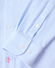 Close-up of the sleeve on the Psycho Bunny MENS BRENDAN JERSEY SHIRT - B6C749D200, crafted in a soft light blue pinstripe from premium Pima cotton. It features a neatly buttoned cuff with a striking pink buttonhole accent against smooth white buttons. The shirt is laid flat, showcasing the sleeve's precise stitching and button detailing.