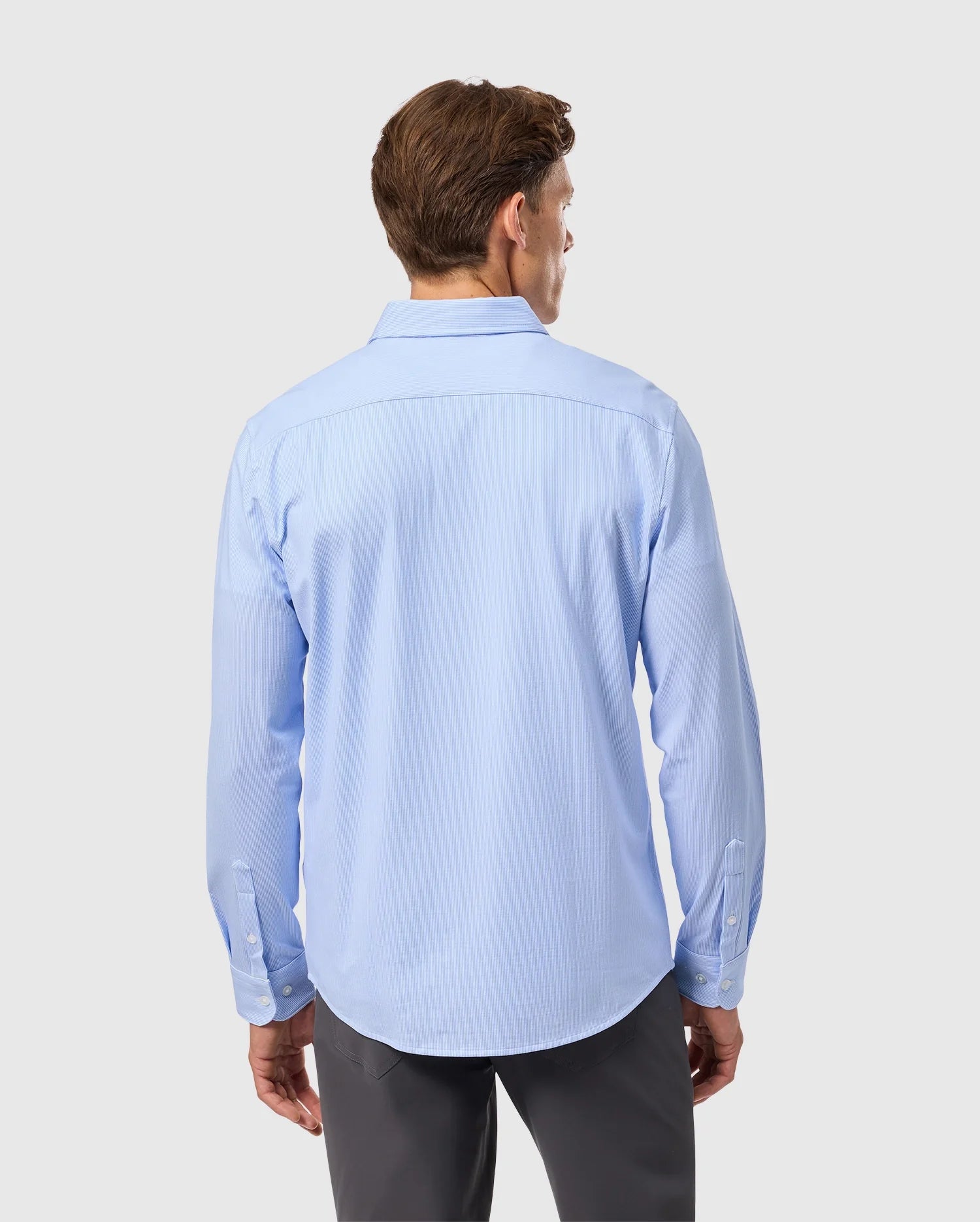 A man wearing a Psycho Bunny light blue striped jersey shirt made of Pima cotton, identified as product B6C750D200, with long sleeves and gray pants, stands facing away from the camera against a white background.