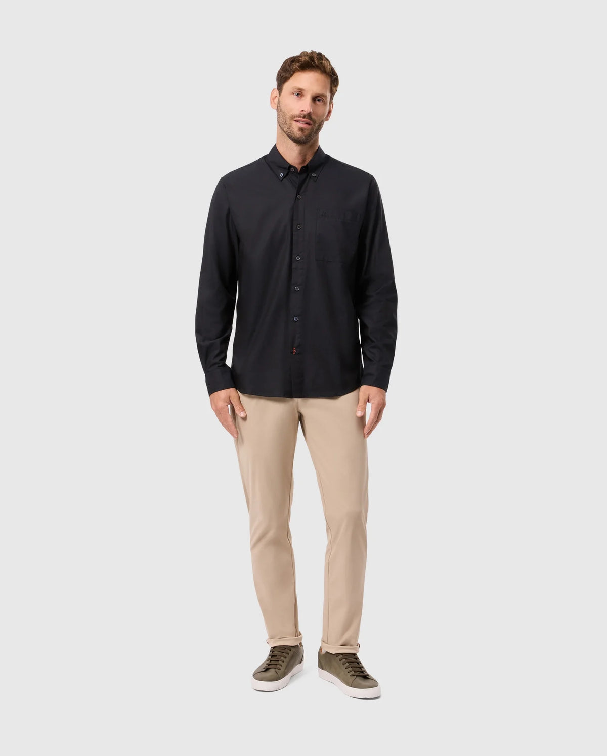 A man stands against a plain background, wearing a Psycho Bunny MENS JARED COTTON MODAL SHIRT in black, beige pants, and green sneakers. His right hand is in his pocket while he maintains a relaxed expression.