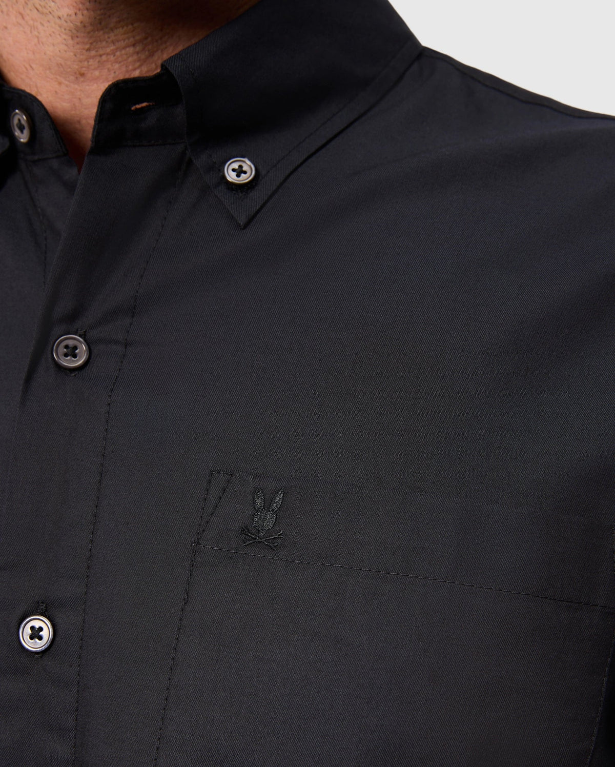 Close-up of a person wearing a Psycho Bunny MENS JARED COTTON MODAL SHIRT - B6C774D200 in black, featuring a small embroidered logo of two overlapping rabbits above the chest pocket. This premium shirt offers style and comfort with matching black buttons and a classic collar.
