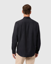 A man is turned away, wearing the Psycho Bunny MENS JARED COTTON MODAL SHIRT - B6C774D200 in black with beige pants, set against a plain gray background.