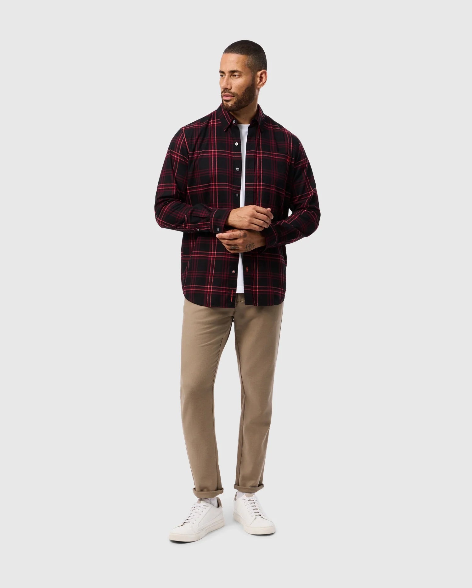 A man stands against a plain background wearing the Psycho Bunny MENS HAYES PLAID SHIRT - B6C842D200, featuring a red and black button-down plaid pattern over a white T-shirt. He pairs this with beige pants and white sneakers. Sporting a beard and short hair, he has his head turned slightly to the side.
