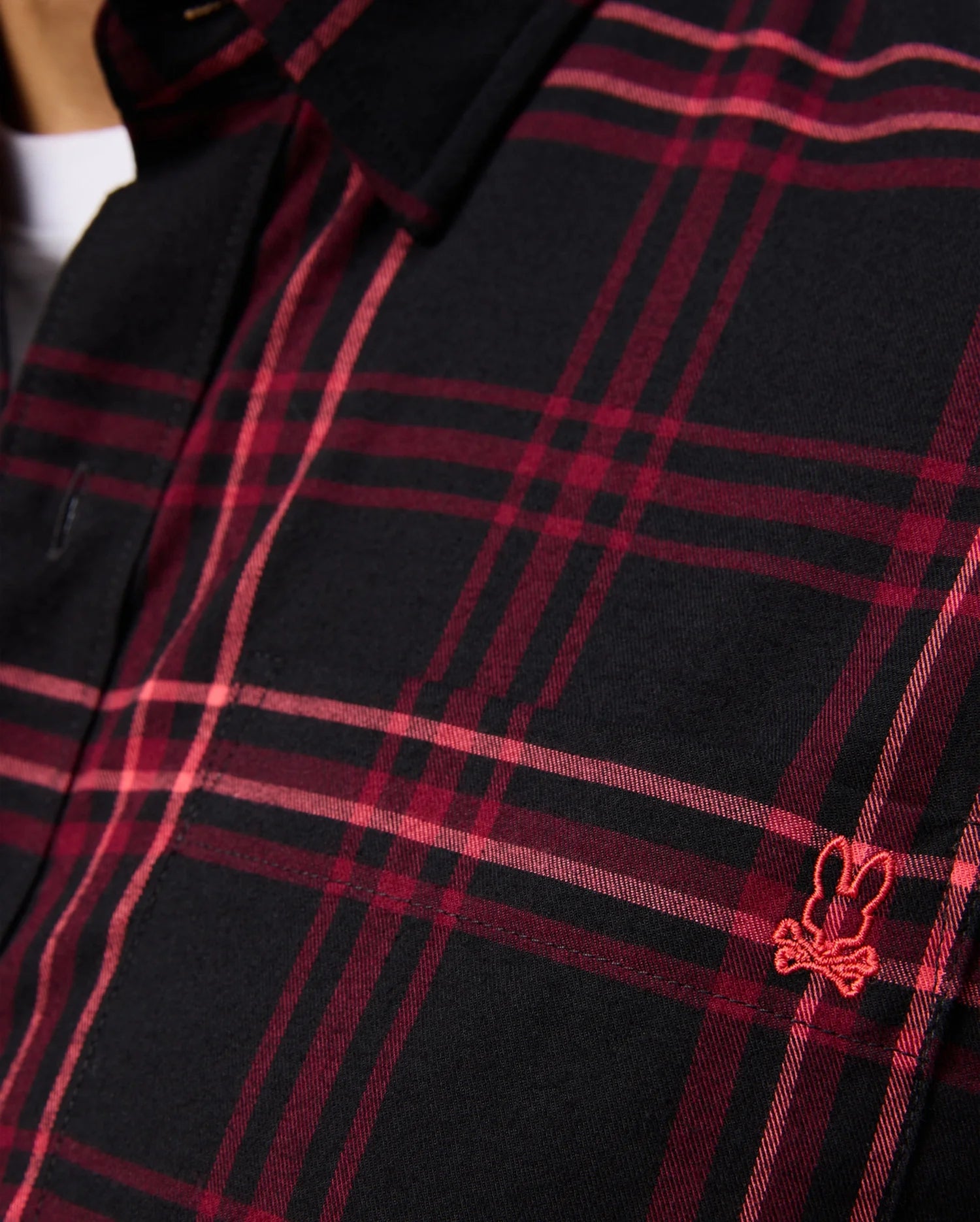 A detailed view of the MENS HAYES PLAID SHIRT - B6C842D200 by Psycho Bunny, showcasing a red plaid design on a black button-down shirt with a small embroidered red bunny logo on the chest, made from soft Pima cotton.