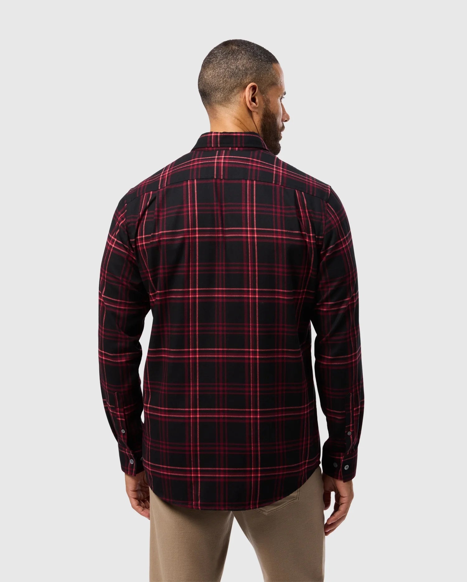 A man is standing with his back to the camera, wearing the Psycho Bunny MENS HAYES PLAID SHIRT (B6C842D200), which features long sleeves and soft Pima cotton fabric. He is also wearing beige pants, set against a plain light gray background.
