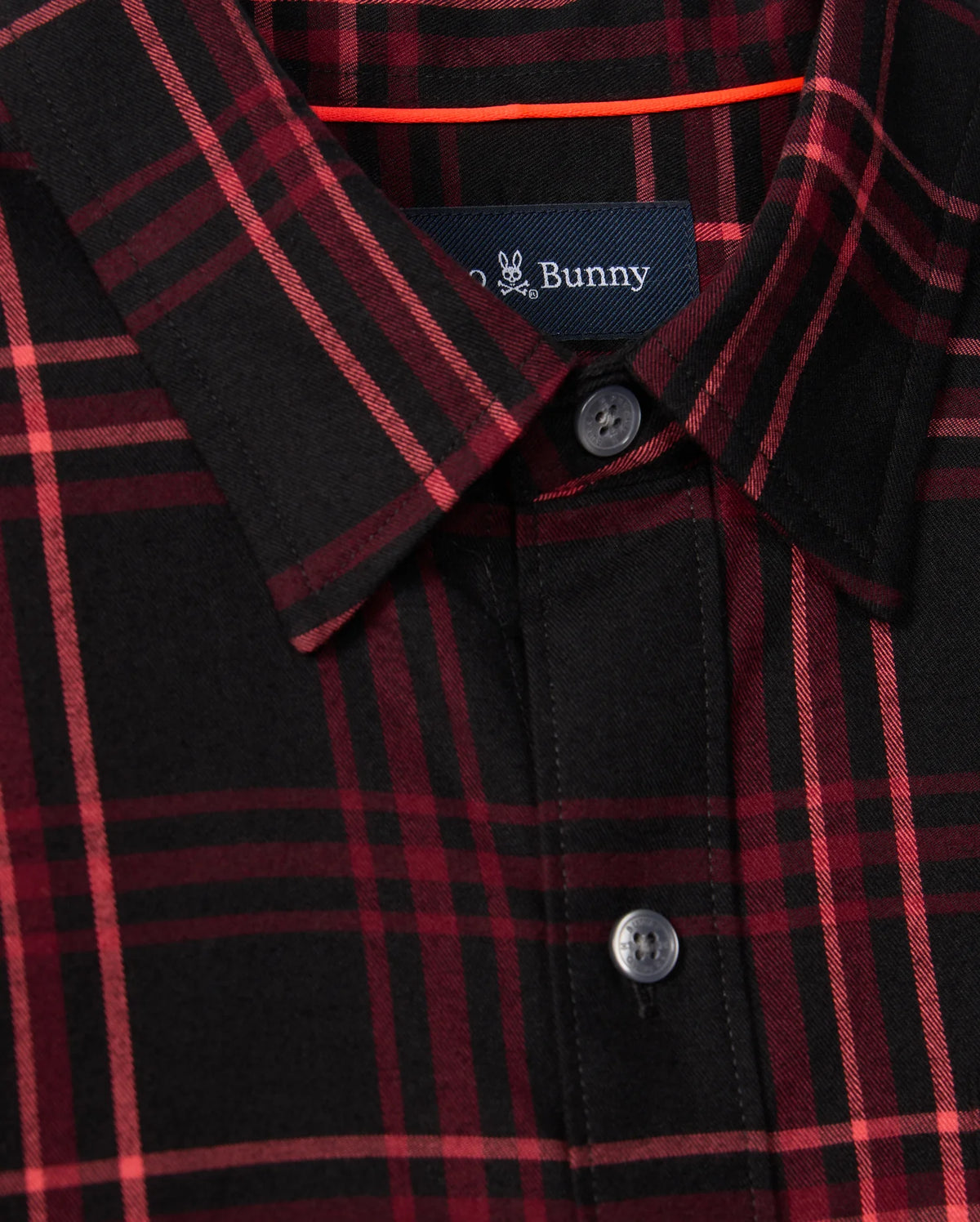 A close-up of the Psycho Bunny MENS HAYES PLAID SHIRT (B6C842D200) showcases its red and black button-down plaid pattern, complemented by a folded collar and visible buttons. Crafted from soft Pima cotton, the shirt includes a small logo label with partially visible text sewn inside the neckline.