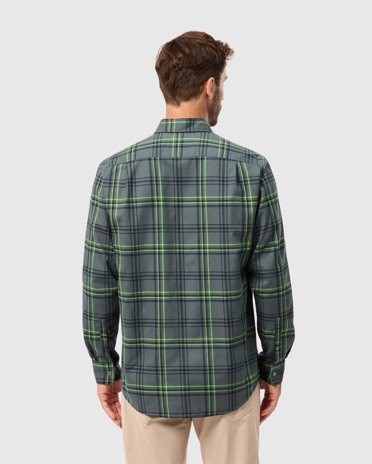 Rear view of a person wearing the Psycho Bunny MENS HAYES PLAID SHIRT - B6C842D200 with long sleeves in green and gray, paired with beige pants. The premium button-down shirt stands out against the plain light gray background.