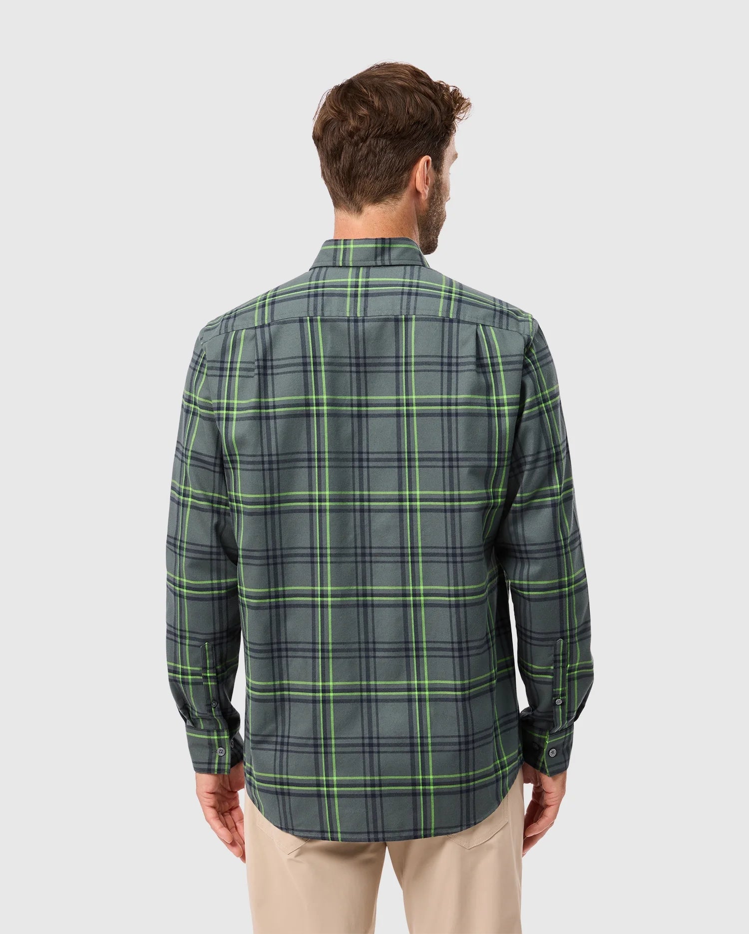 Rear view of a person wearing the Psycho Bunny MENS HAYES PLAID SHIRT - B6C842D200 with long sleeves in green and gray, paired with beige pants. The premium button-down shirt stands out against the plain light gray background.
