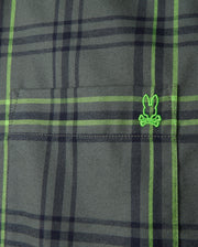 Close-up of the Psycho Bunny Men's Hayes Plaid Shirt - B6C842D200, crafted from premium Pima cotton. The green fabric, featuring intersecting black lines, showcases a small embroidered logo resembling a cartoonish face wearing glasses. The texture and stitching detail of this button-down shirt are clearly visible.