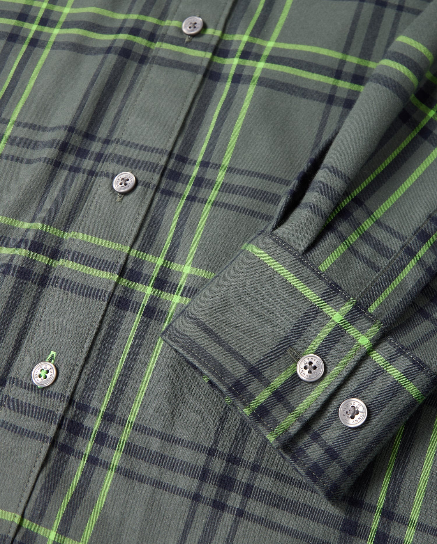 Close-up of the MENS HAYES PLAID SHIRT - B6C842D200 by Psycho Bunny, featuring a pattern of green and dark blue lines. The Pima cotton fabric is neatly folded, displaying a buttoned sleeve cuff and several visible buttons down the front. The material exudes a soft and casually elegant appearance.
