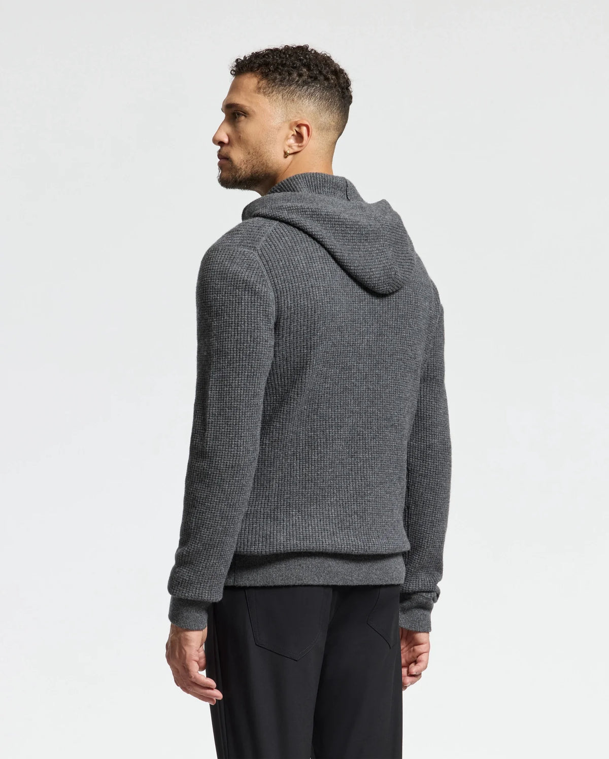 A person with short curly hair and a beard is facing left, wearing a grey Psycho Bunny MENS LEVAN WAFFLE HOODED SWEATER with an embroidered chest bunny and black pants against a plain light background.