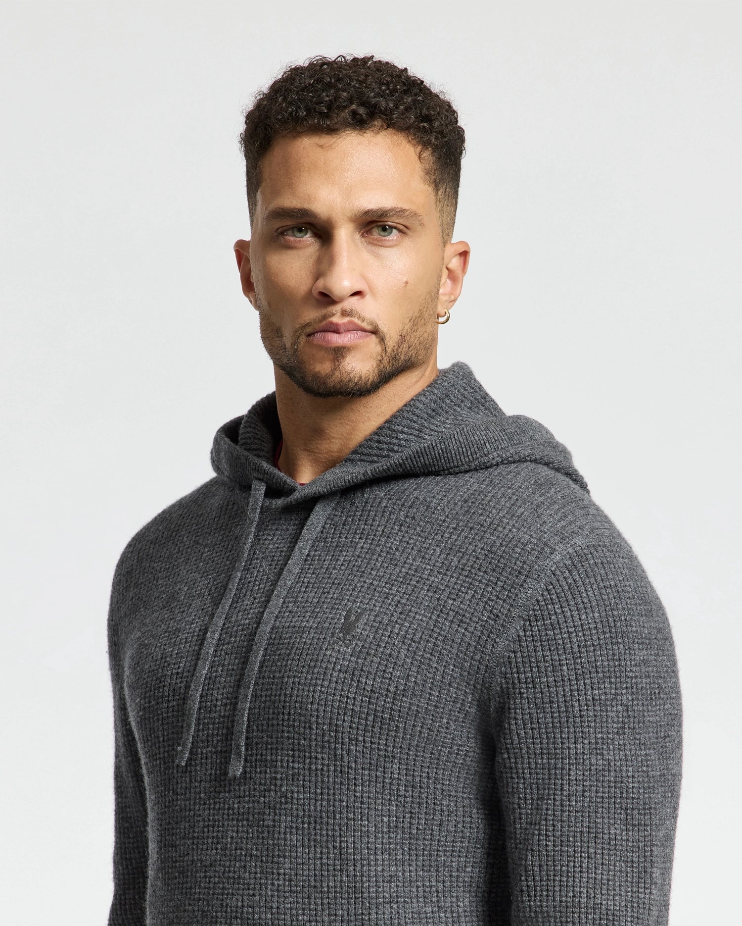 A man with short curly hair wears a Psycho Bunny MENS LEVAN WAFFLE HOODED SWEATER (B6E246E200). He stands against a plain light background, his serious expression complemented by an embroidered chest bunny.