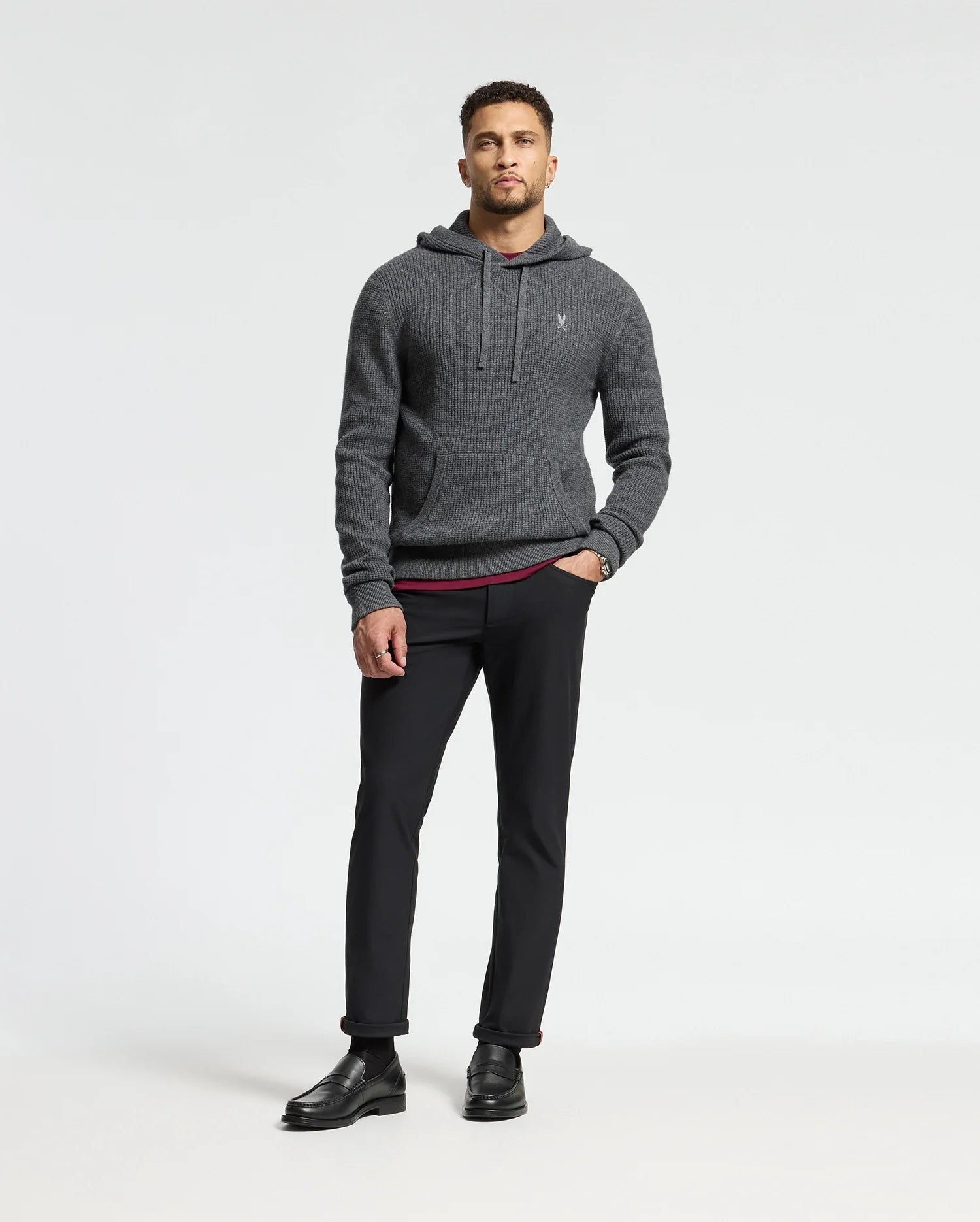 A man in a Psycho Bunny dark gray MENS LEVAN WAFFLE HOODED SWEATER, black pants, and shoes stands against a plain white background. He has short hair and is looking directly at the camera.