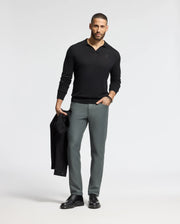 A man with short hair and a beard stands against a plain background, wearing the sleek black MENS JOHNNY COLLAR SWEATER - B6E258E200 by Psycho Bunny and gray pants. He holds a black jacket in one hand, complementing his classic look with black shoes.