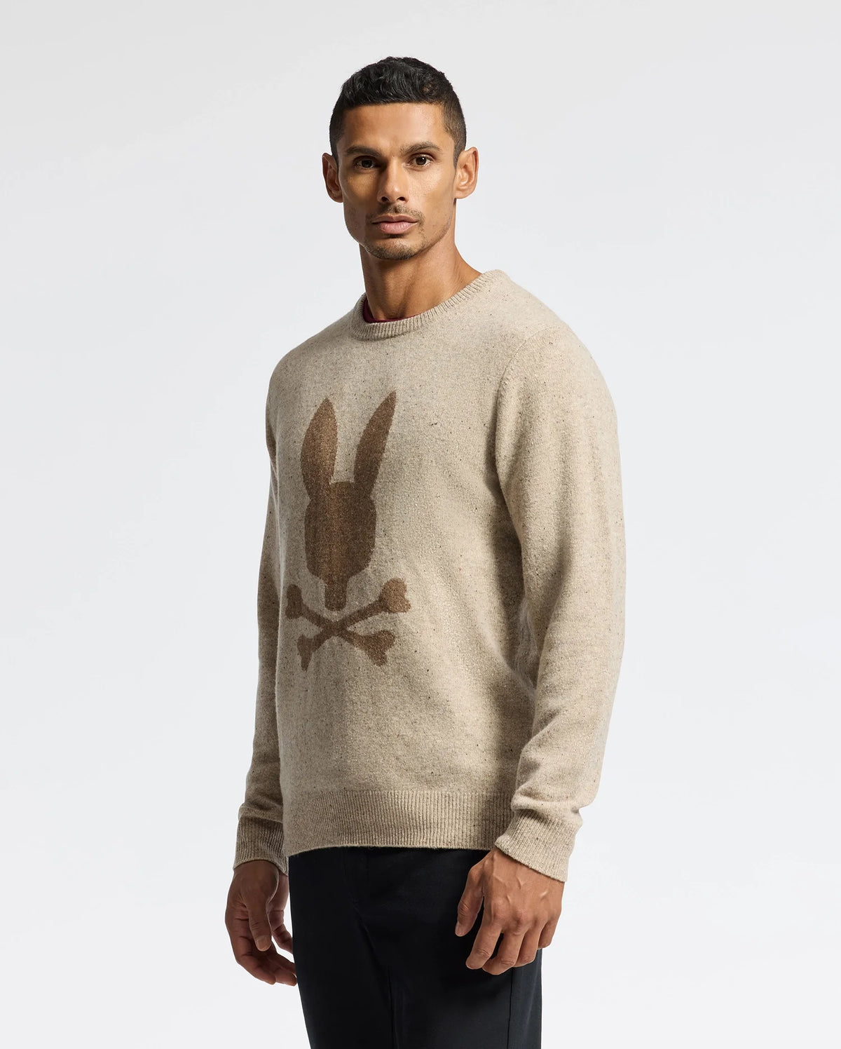 A man wearing the MENS LINCOLN MERINO WOOL SWEATER by Psycho Bunny, featuring a brown logo of bunny ears and crossed bones, stands against a plain white background, gazing directly at the camera.